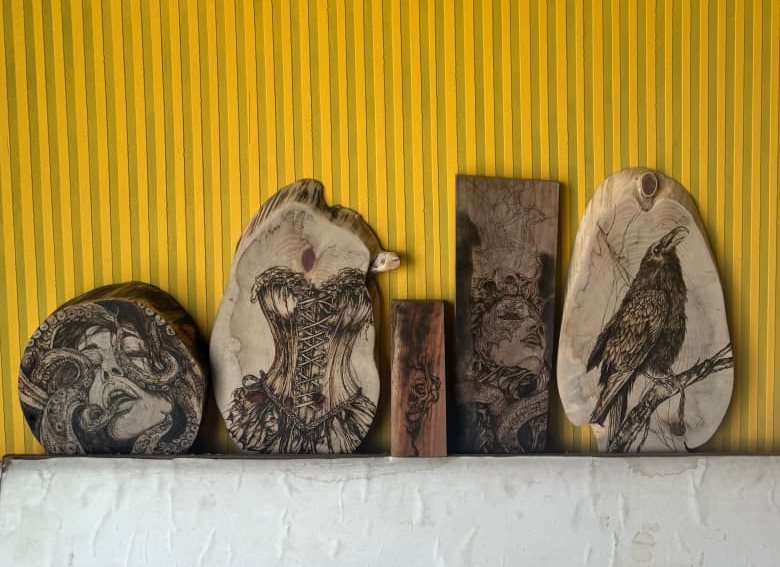 Five new pyrography works leaning against a yellow striped wall: a woman with octopus tentacles in her hair on a round slab, a corset on a shaped slab, a skull study on a narrow plank, a serpent-wreathed face on a dark panel, and a raven perched on a branch on a live-edge oval