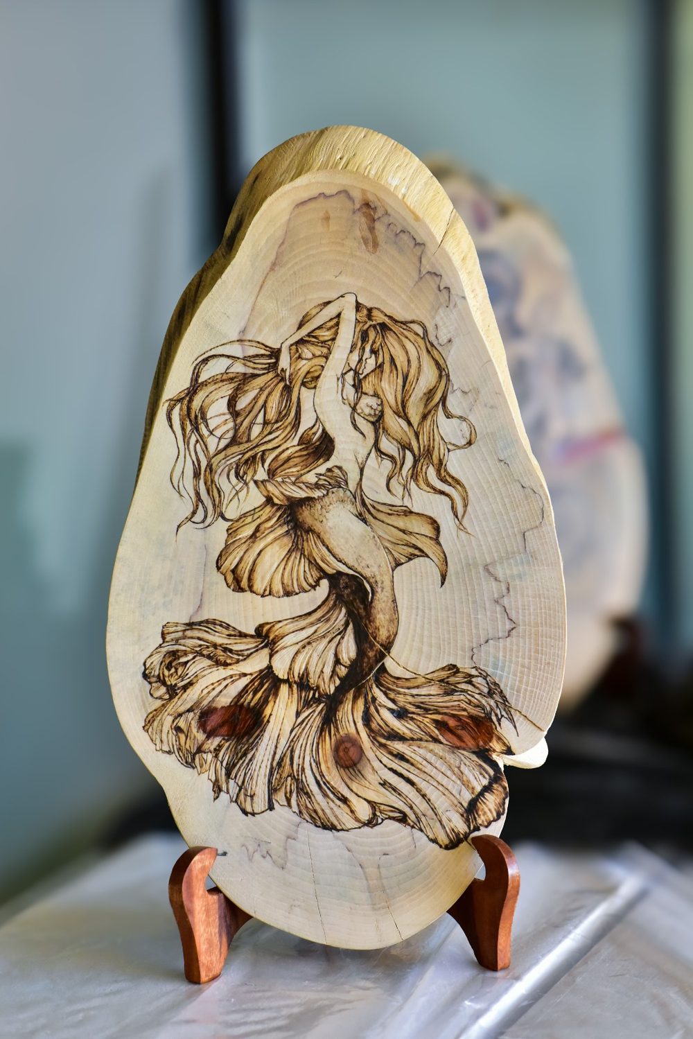 The Siren's Call — a mermaid with flowing hair and a fin-like tail, burned on an oval live-edge slab standing on a wooden easel