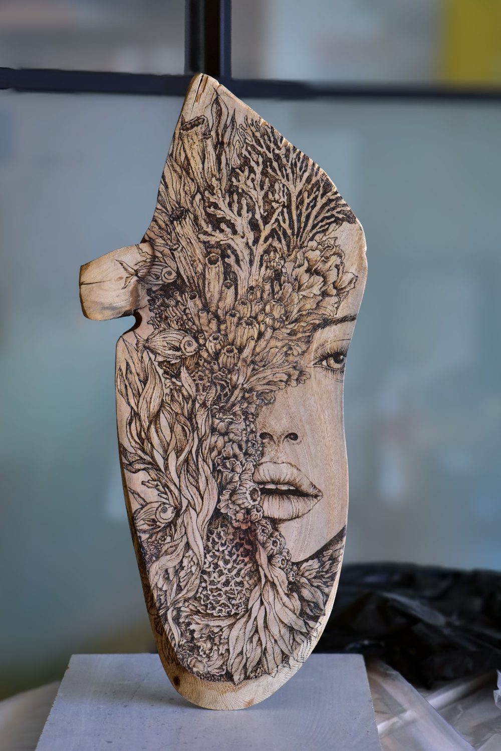Still Becoming — a woman's face emerging from burned coral, kelp and reef life on a tall, free-standing live-edge wood slab